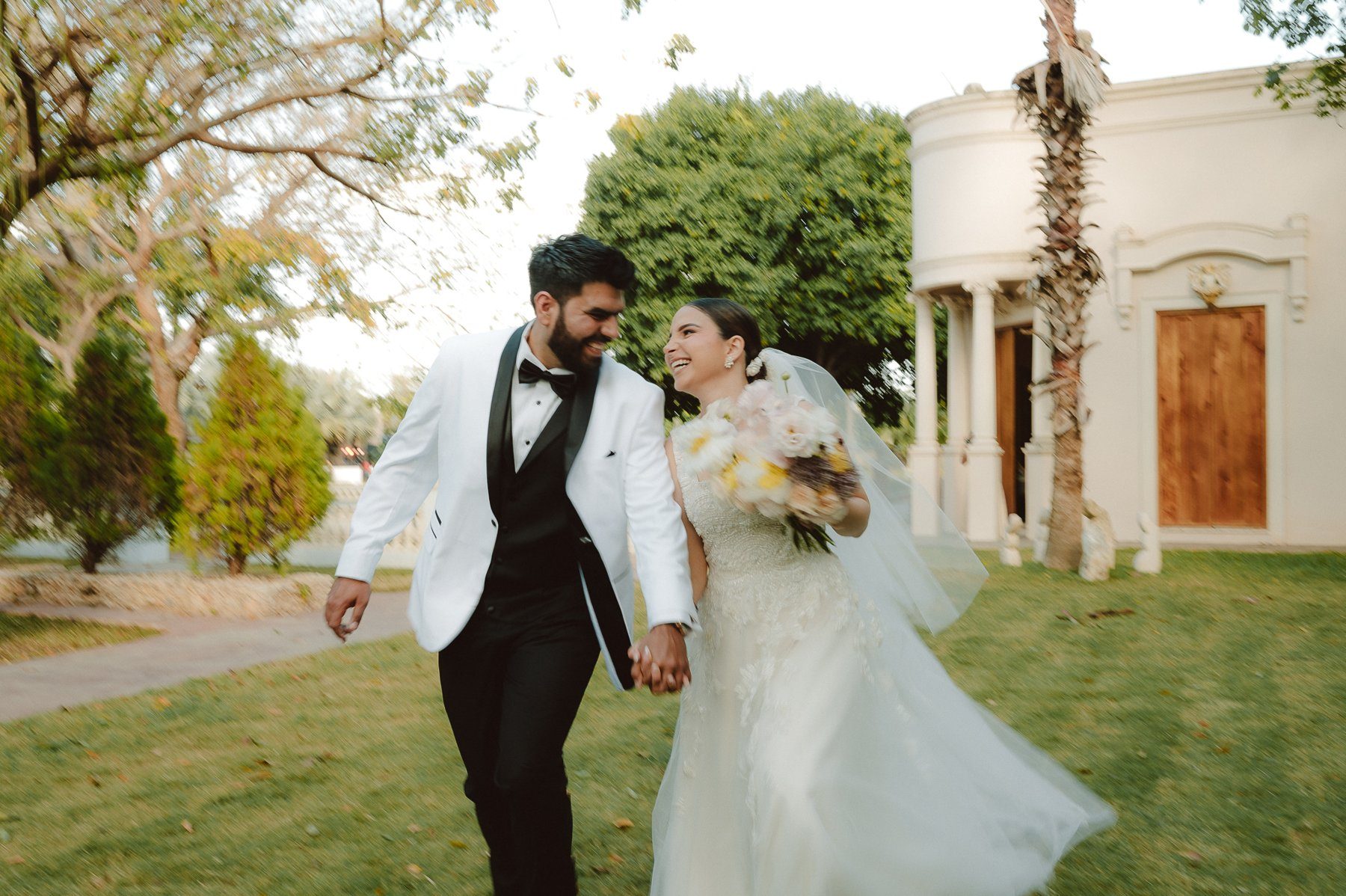 Pareja de novios riendo mientras caminan de la mano por el jardín de una hacienda